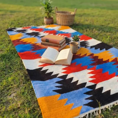 Colorful handwoven shataranji rug on green grass with books, coffee mug and small plants – perfect for picnic and cozy reading corner | সবুজ ঘাসের ওপর বিছানো রঙিন হাতে বোনা শতরঞ্জি, ওপরে বই, কফির মগ আর ছোট গাছ রাখা – পিকনিক ও রিডিং কর্নারের জন্য উপযুক্ত