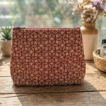A handcrafted jute cosmetic pouch with a red floral pattern, displayed on a wooden table with plants and natural skincare items in the background.