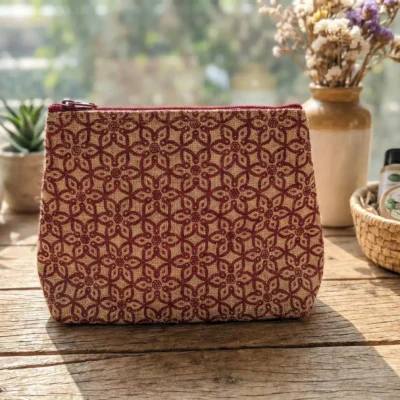 A handcrafted jute cosmetic pouch with a red floral pattern, displayed on a wooden table with plants and natural skincare items in the background.